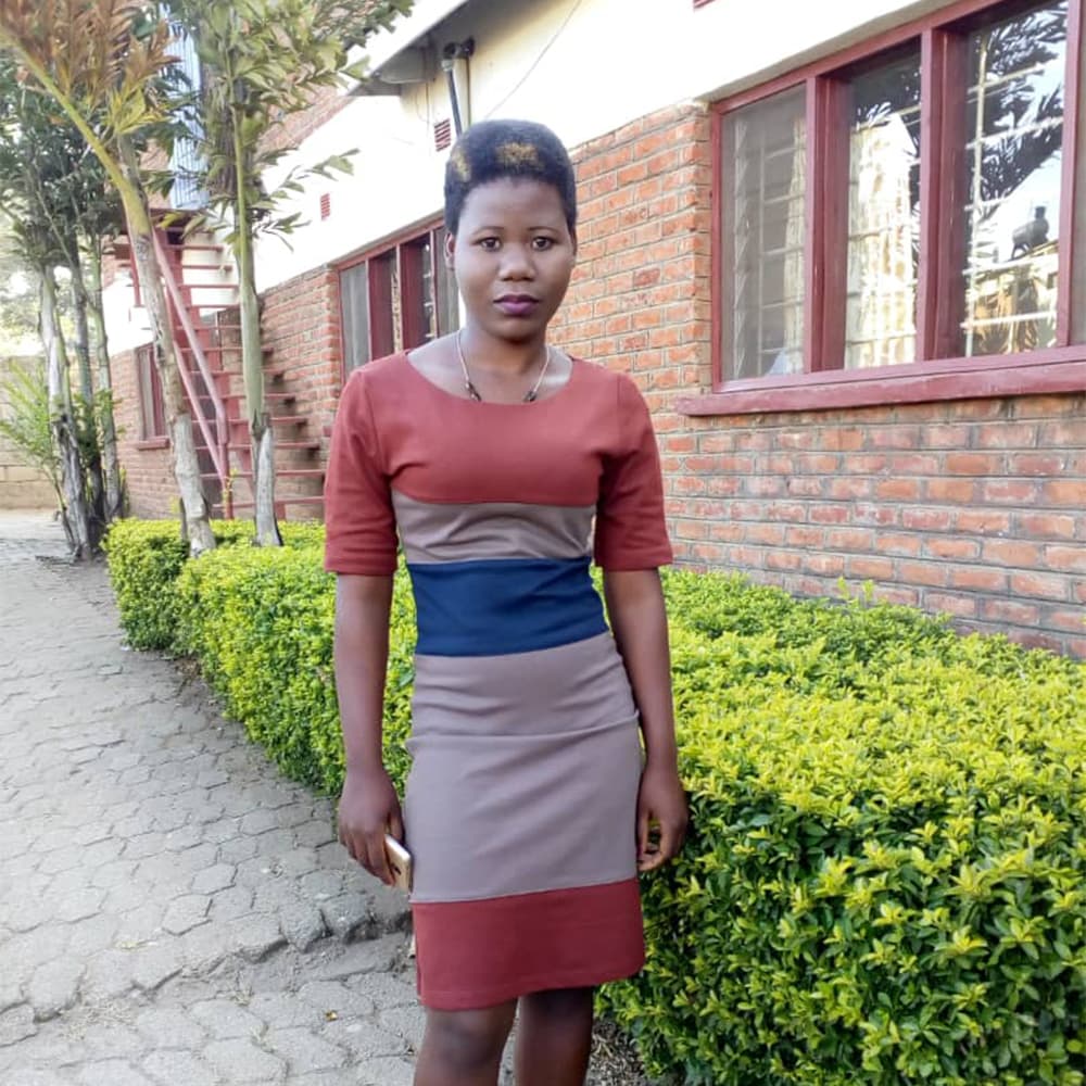 Malawi-1---Chimwemwe A young woman in Malawi, with a short afro hairstyle and wearing a striped red and blue dress, looking at the camera while standing outside of a red brick building surrounded by bright green shrubs.