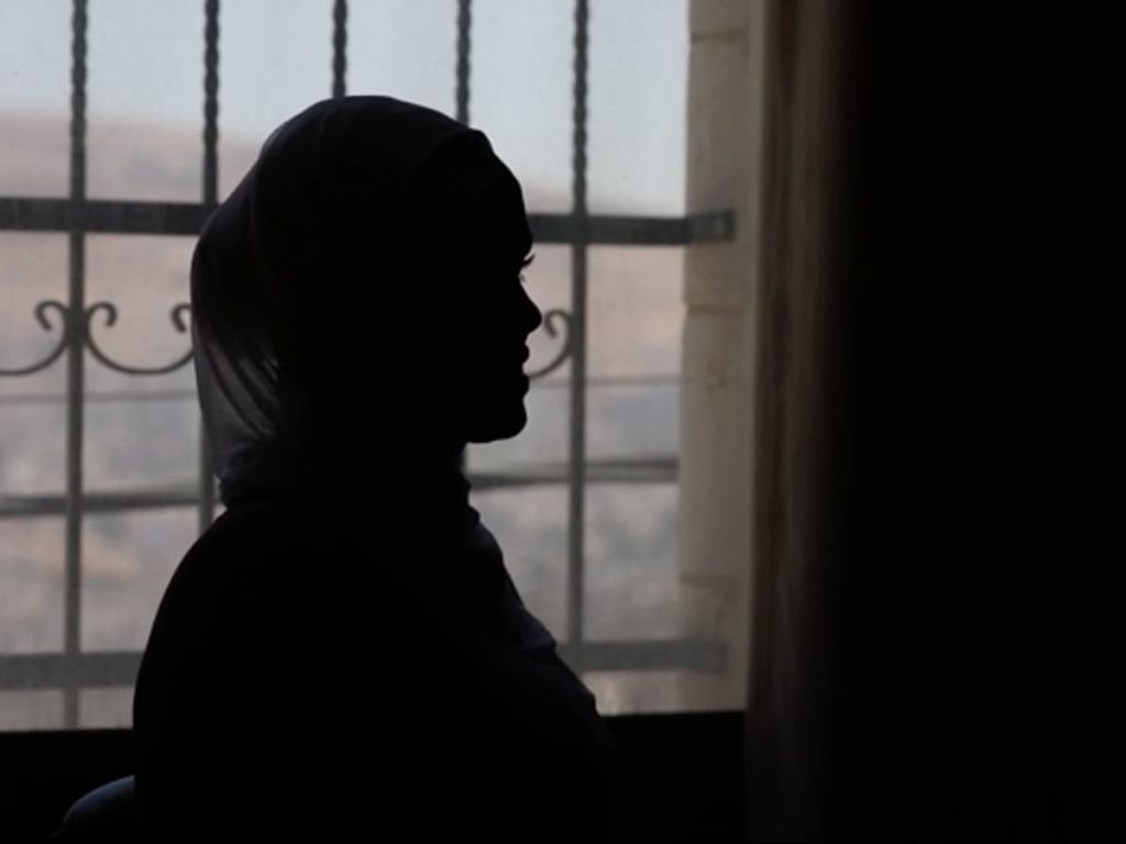 A silhouette of a Palestinian woman in front of a barred window in a dark room.