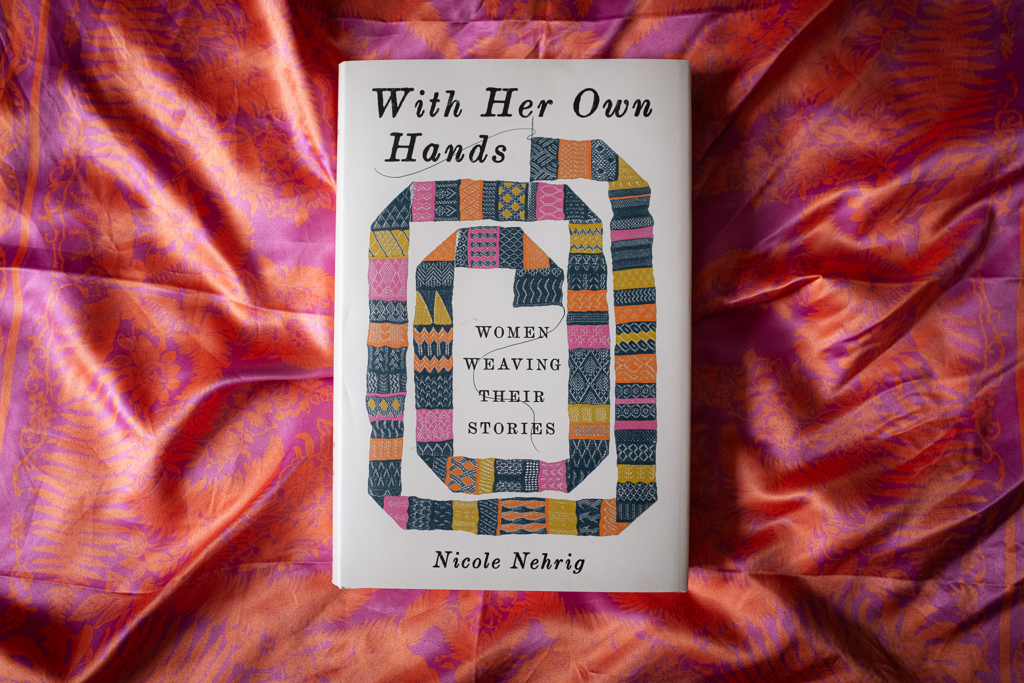 Women Weaving Their Own Stories, a book by Nicole Nehrig, lies flat on a pink satin fabric.