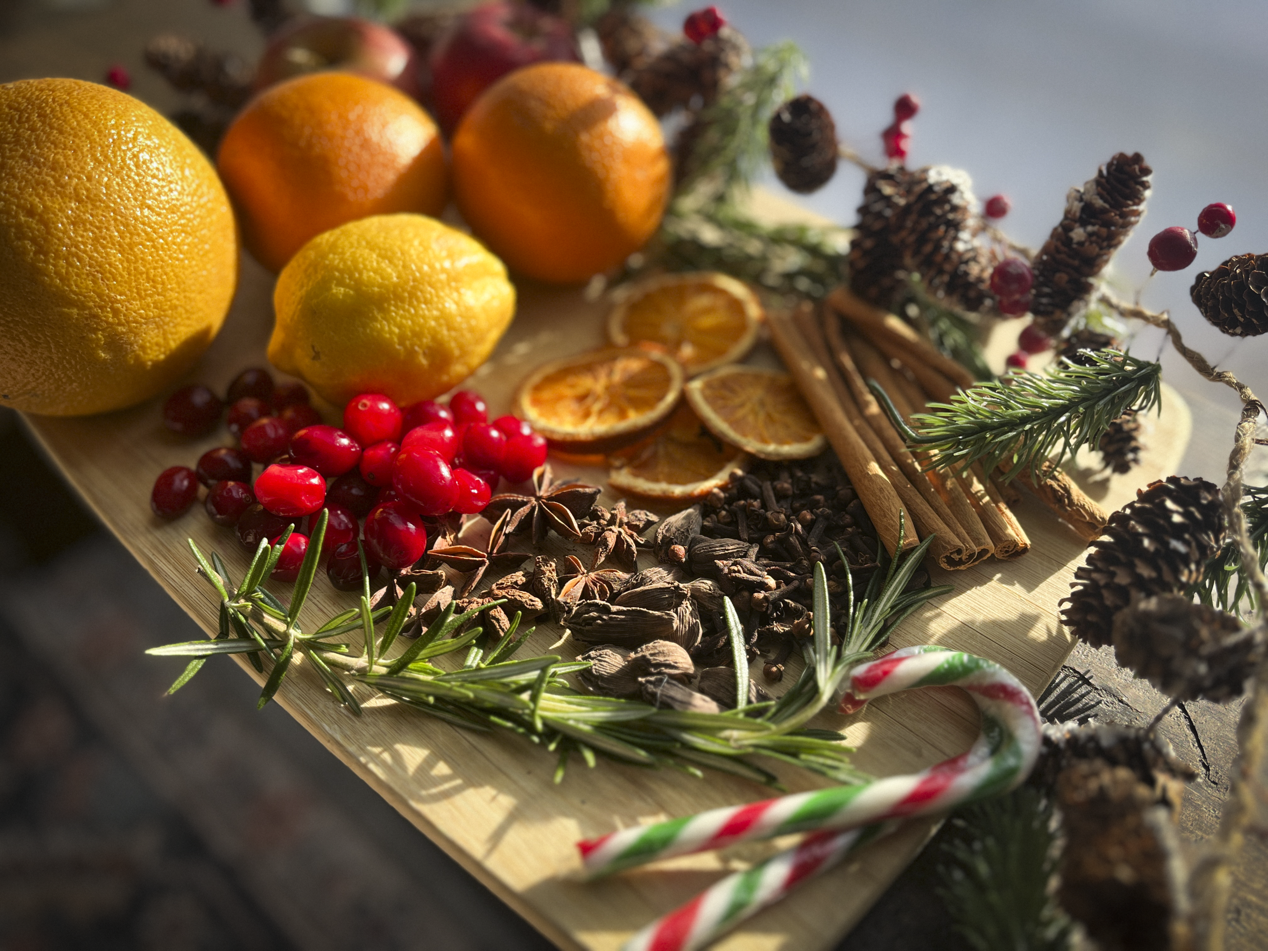 Various simmer-pot ingredients arranged on a wooden tray, including oranges, lemons, cranberries, cinnamon sticks, star anise, and rosemary.
