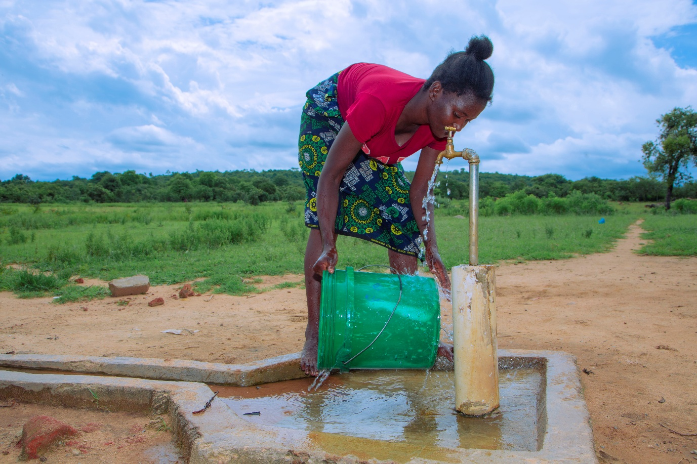 A woman wearing a red shirt, and a green and black skirt washing a bucket at a borehole.