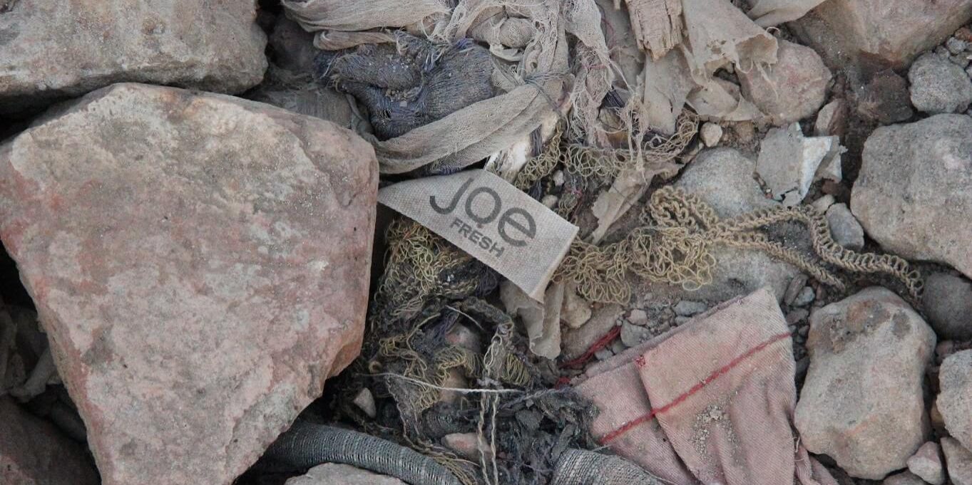 Joe-Fresh-stock-image Grey rocks of varying sizes along with parts of grey torn cotton material are mixed with pieces of old wood and string. A dirty Joe Fresh label sits in the middle of the rocks on the ground.