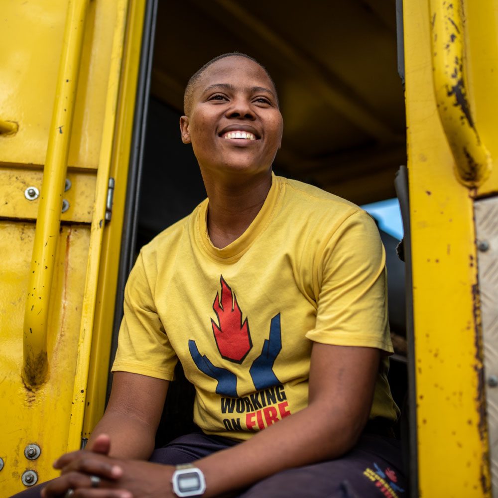 A female firefighter sits in a firetruck, smiling and looking away from the camera.