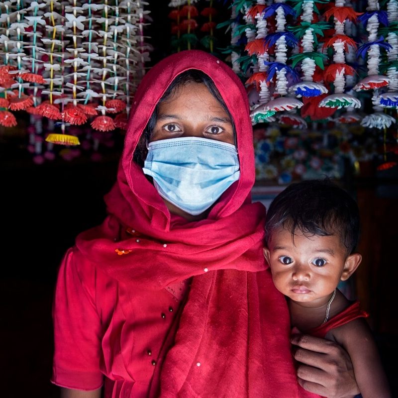 A woman with brown skin, brown eyes and dark hair is holding her young child and wearing a blue surgical mask and a red dress and headscarf.
