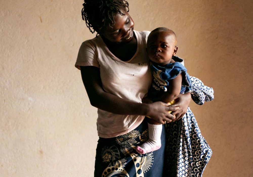 SexualReproductiveRights_Hero1 A young woman stands, smiles at her infant son she is holding in her arms while he looks at the camera.