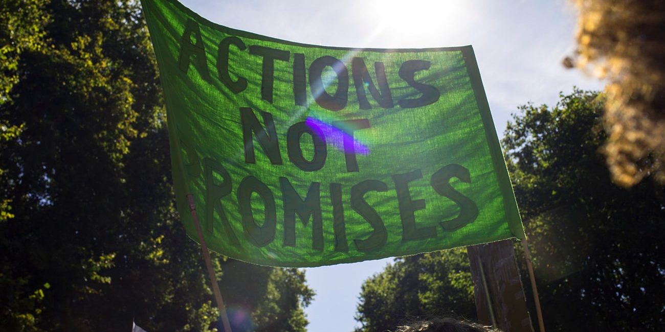 A green banner being held up at a protest that says : Actions Not Promises