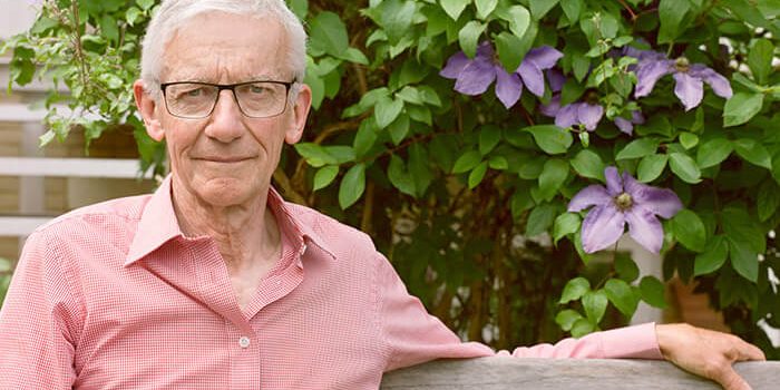 An elderly man wearing glasses and a light-red shirt, sits in a bench outdoors with his left hand stretched out looking firmly at us.