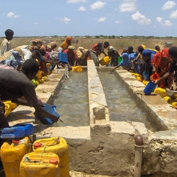 A large group of dark-skinned people of various genders and ages are gathering around a large, above ground, stone water source, filling large yellow jerry cans with water.