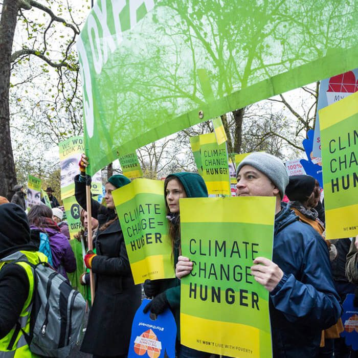 A group of mostly light-skinned people of varying genders and ages wearing winter jackets, hoodies, hat and down vests are gathered in protest of climate change. They are holding identical protest signs that read Climate Change Hunger in white letters on a multi-toned green striped background. There is a serious mood to the photo.