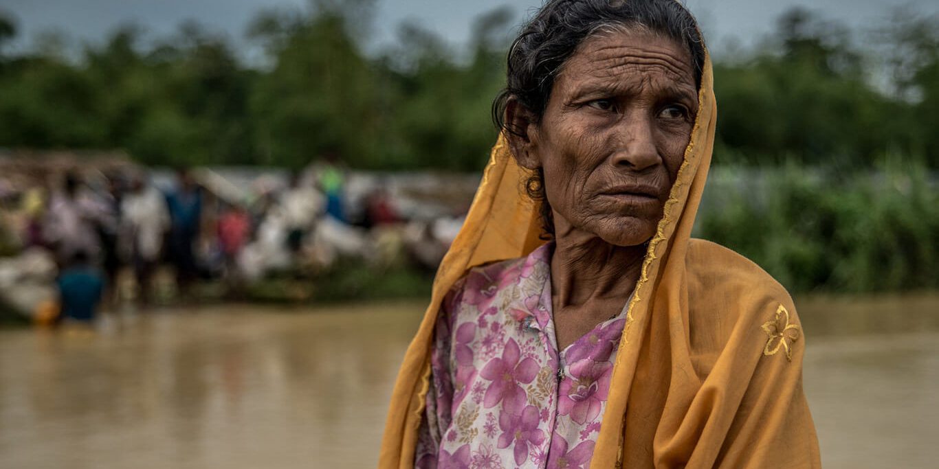 An aging, brown-skinned Rohingya woman with black hair and brown eyes is wearing a pink and white floral button-down dress and a mustard yellow scarf over her head. She stands by the river with a serious, almost pained look on her face. On the other side of the river, there is a crowd of people gathered near the river's edge. It's not clear what they're doing because there is a blurred effect on the background of the photo.