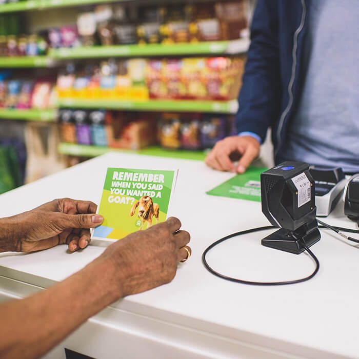 A brown-skinned person whose hands are the only things that are visible in the photo holds a card with a picture of a goat on it that reads Remember when you said you wanted a goat? The person is standing at a white retail counter. Across the counter stands a light-skinned person whose torso and arms can only be seen. Behind them are shelves lined with various consumer products.